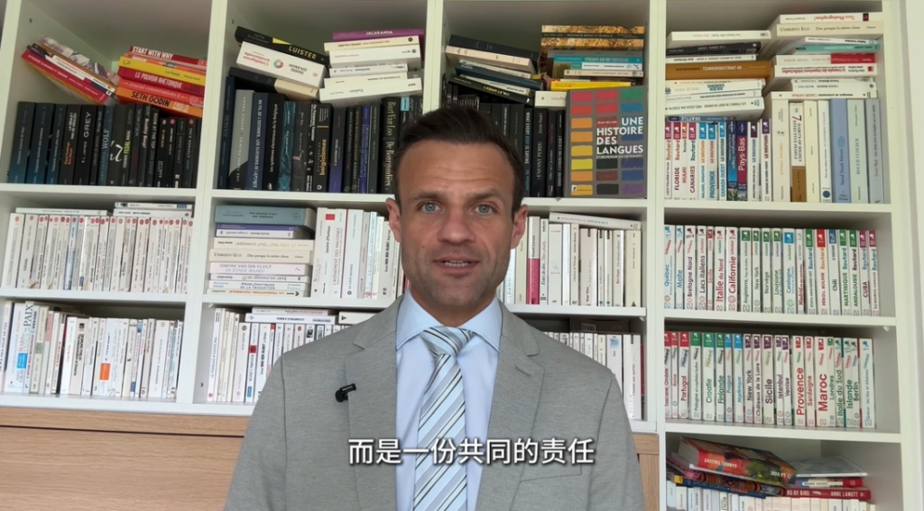 a man addressing a camera with a large bookcase behind him